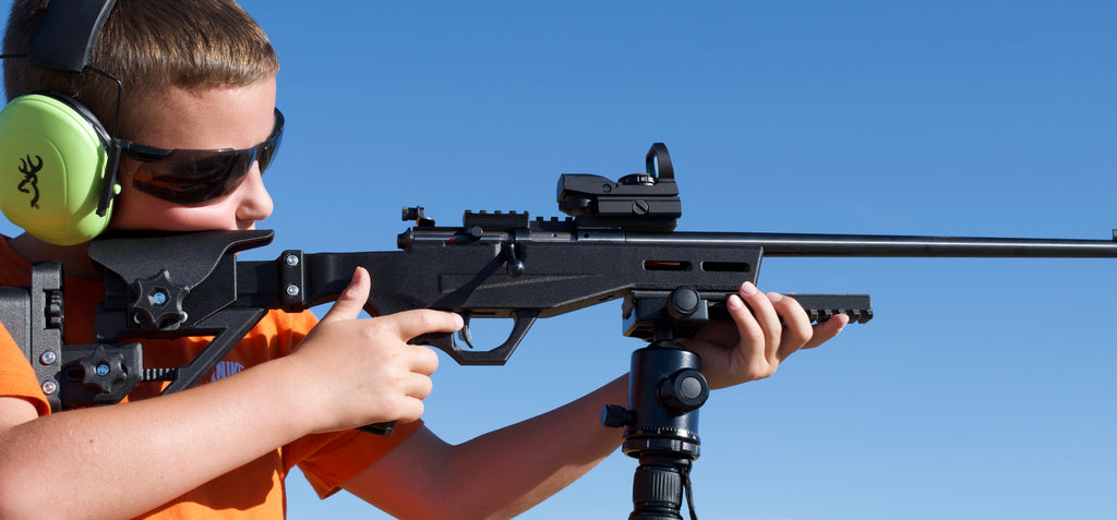 A young boy wearing protective ear muffs and sunglasses aims a black rifle with a scope and bipod against a clear blue sky.
