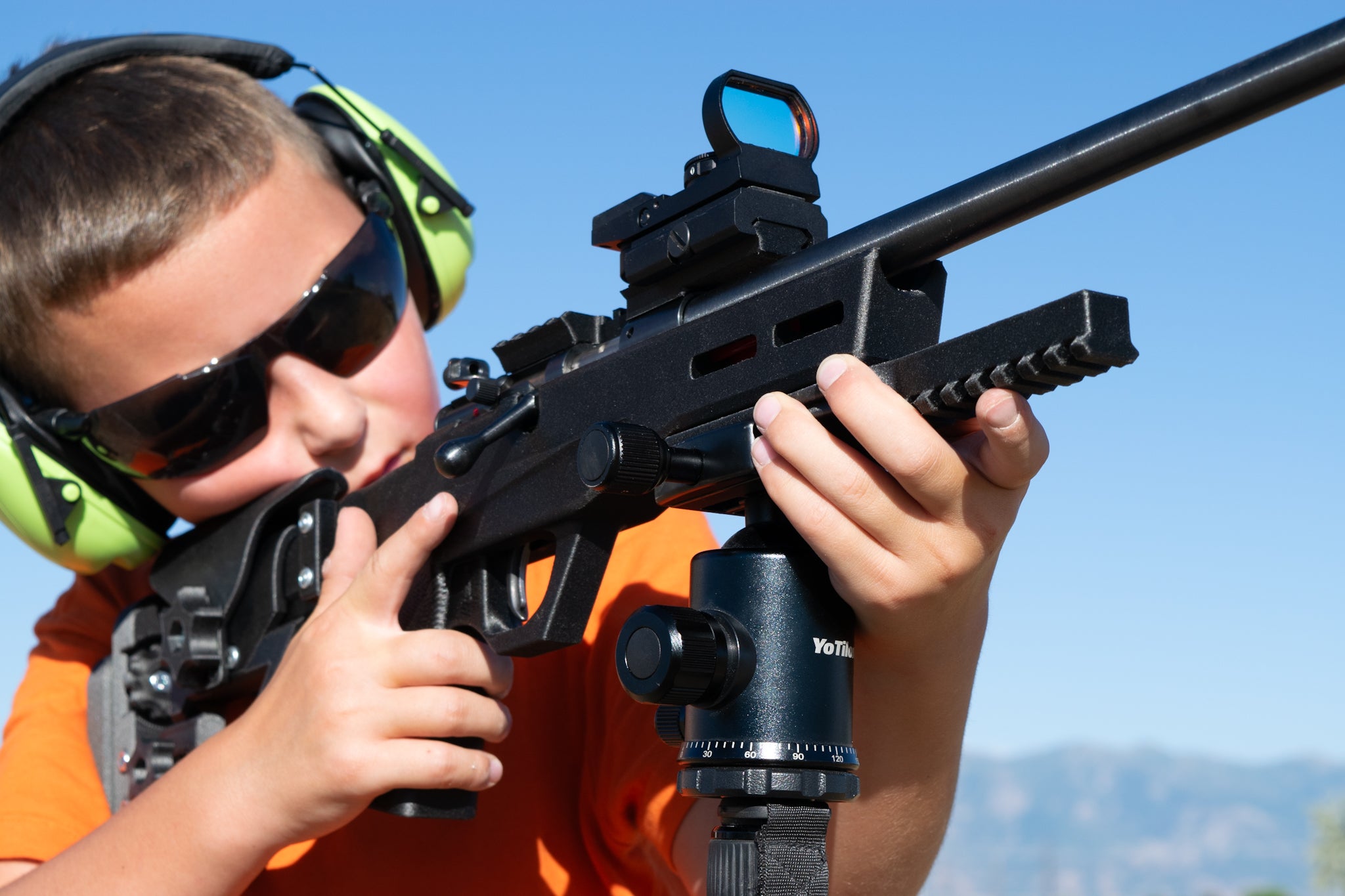 A person wearing protective eyewear and bright green ear muffs aims a black rifle with a red dot sight mounted on a bipod outdoors under a clear blue sky.