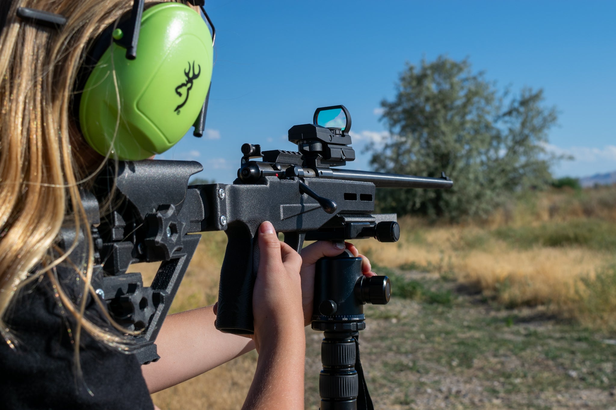 A person wearing bright lime green ear protection aims a black rifle with a red dot sight mounted on a bipod in an outdoor field under a blue sky.