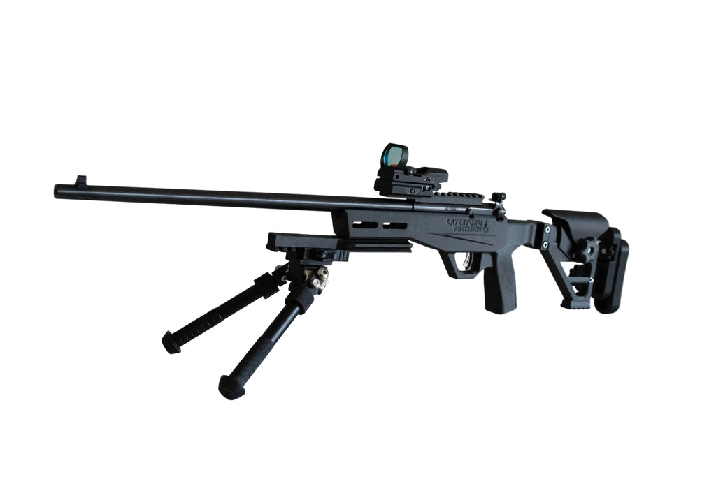 A black tactical sniper rifle with a bipod and red dot sight, resting on a white reflective surface against a beige wall.