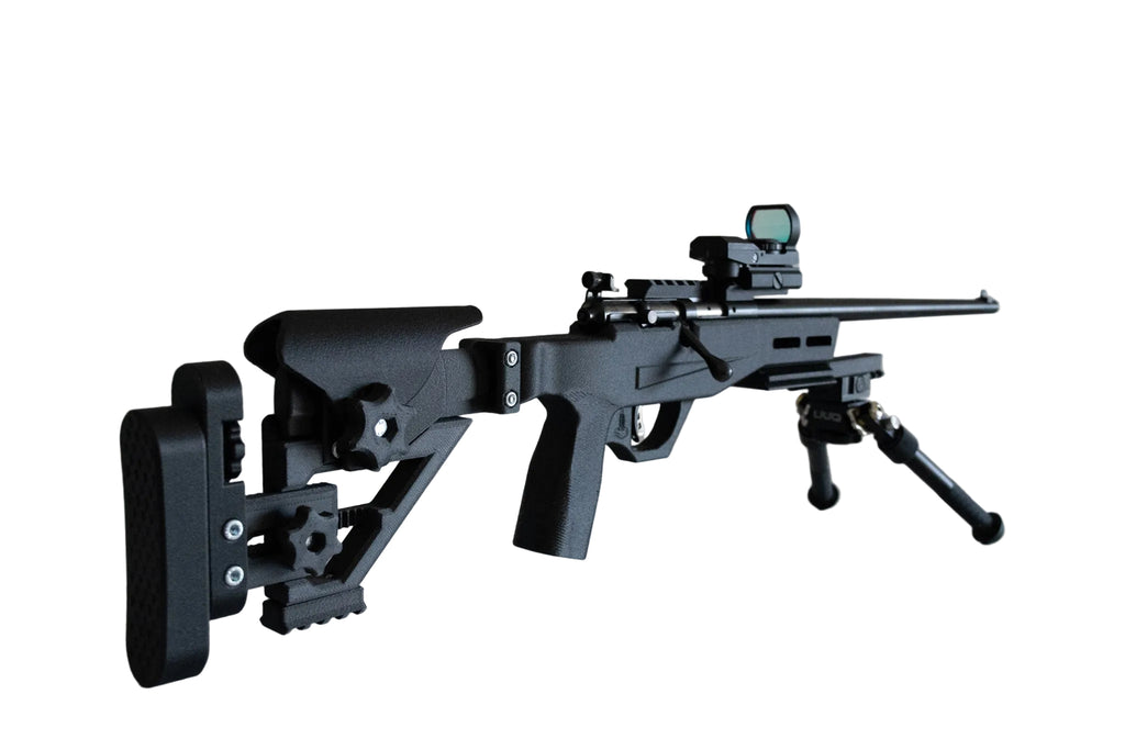 A black tactical sniper rifle replica or model shown on a white reflective surface against a plain beige wall, equipped with a bipod, scope, and adjustable stock.
