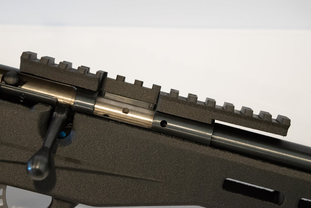 Close-up of a black tactical rifle stock with a mounted black rail system and metallic bolt carrier visible, photographed against a plain light background.