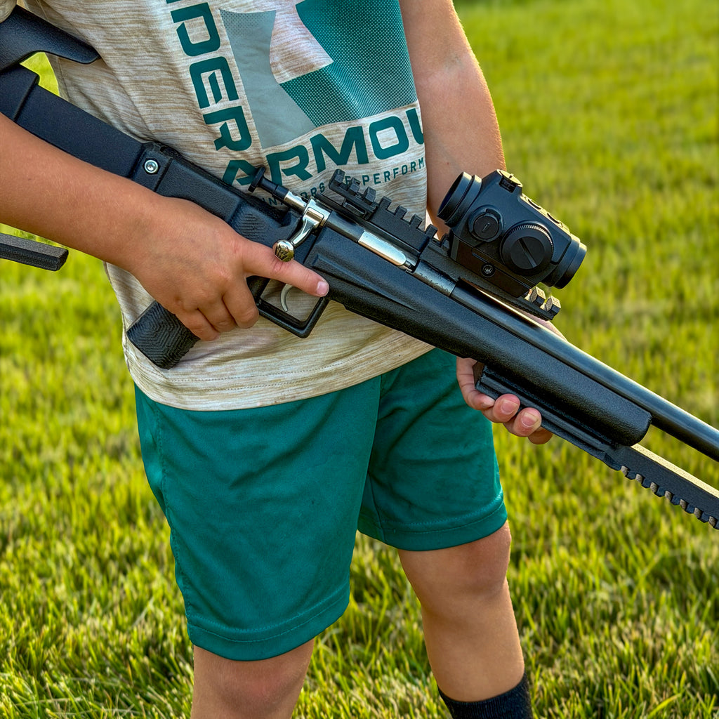 A person outdoors on grass holding a black bolt-action style rifle with a mounted scope. The person wears a light heather gray t-shirt with green lettering and teal-green shorts.