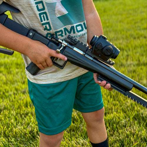 A person outdoors on grass holding a black bolt-action style rifle with a mounted scope. The person wears a light heather gray t-shirt with green lettering and teal-green shorts.