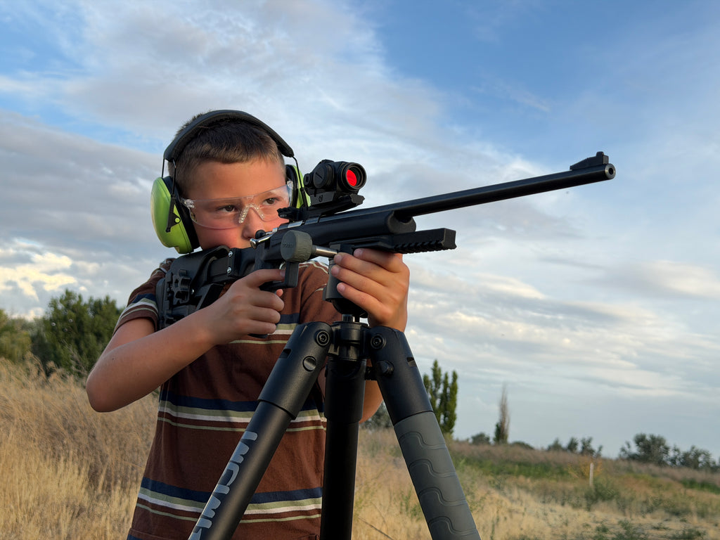 A young boy wearing protective ear muffs and safety glasses aims a black rifle with a scope mounted on a tripod in an outdoor field under a cloudy blue sky.