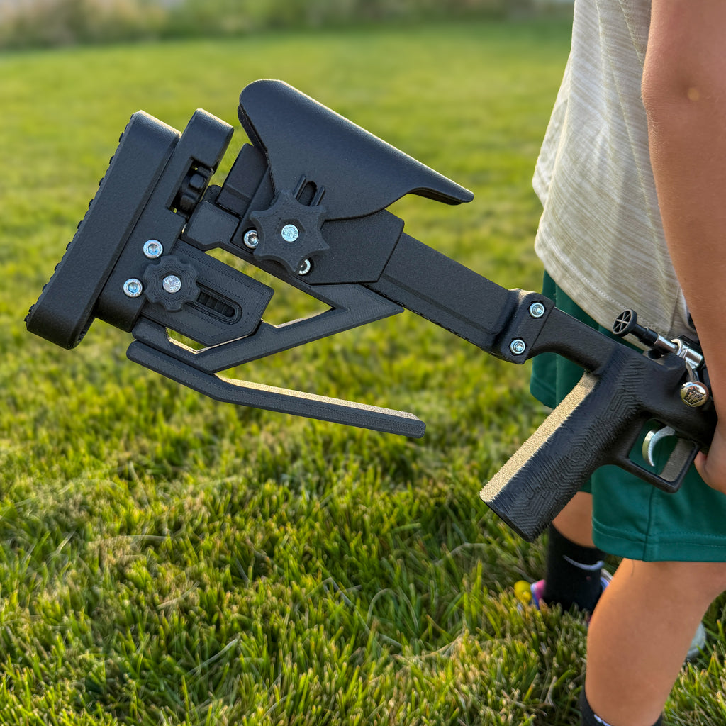 A close-up outdoor shot of a person holding a black adjustable firearm stock and handguard in grass.