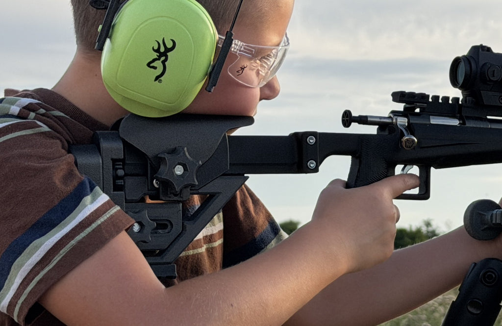 A person wearing protective eyewear and bright green ear muffs aims a black rifle with a scope outdoors. The ear muffs show a black Browning buckmark logo, indicating the brand is Browning.