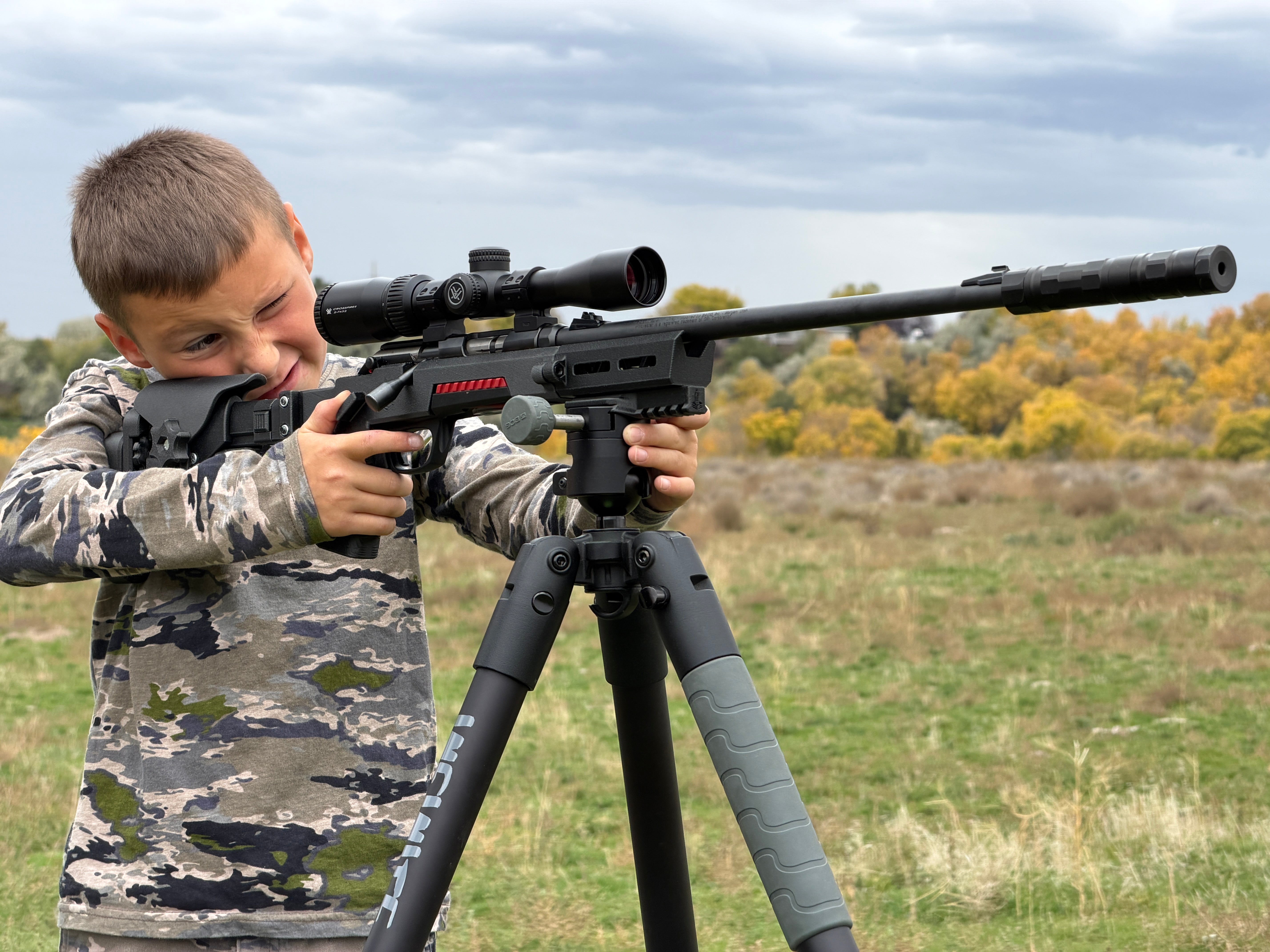 A boy in a digital camouflage long-sleeve shirt aims a black rifle with a scope and suppressor mounted on a black tripod in an outdoor grassy field under a cloudy sky.