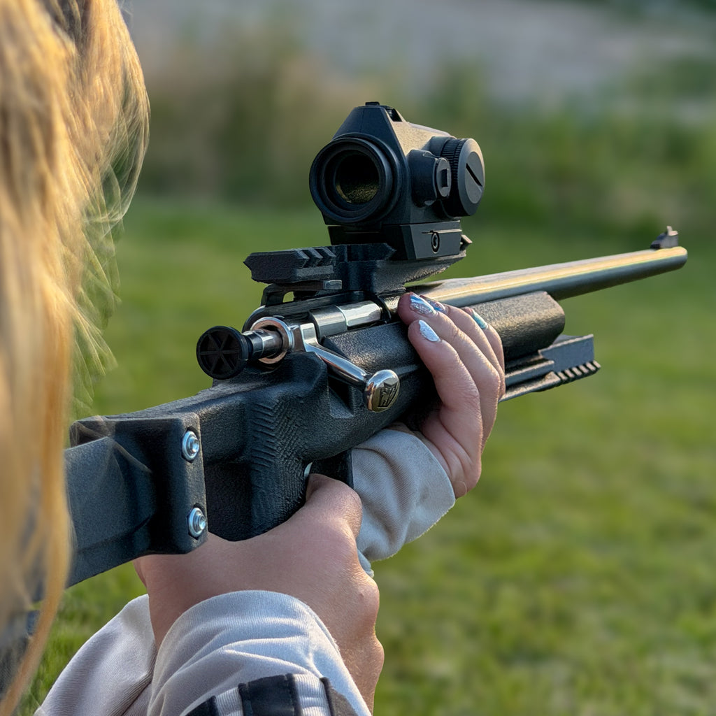 A close-up lifestyle shot of a person aiming a black tactical rifle with an attached scope in an outdoor grassy field.