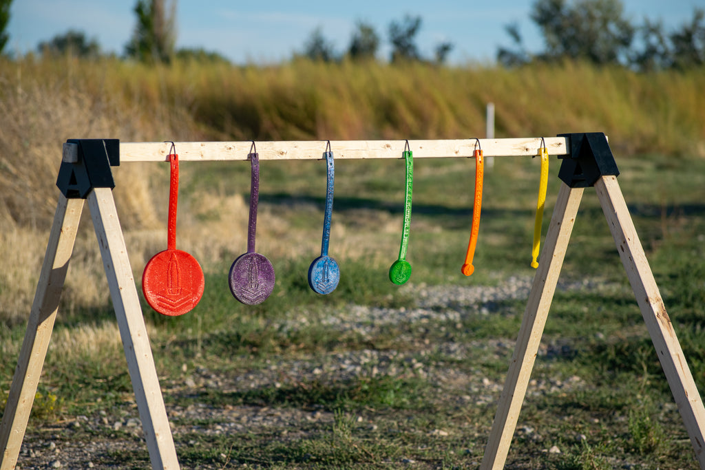 A wooden A-frame stand outdoors with six colorful hanging circular targets in red, purple, blue, green, orange, and yellow, set against a blurred grassy field background.
