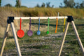 A wooden A-frame stand outdoors with six colorful hanging circular targets in red, purple, blue, green, orange, and yellow, set against a blurred grassy field background.