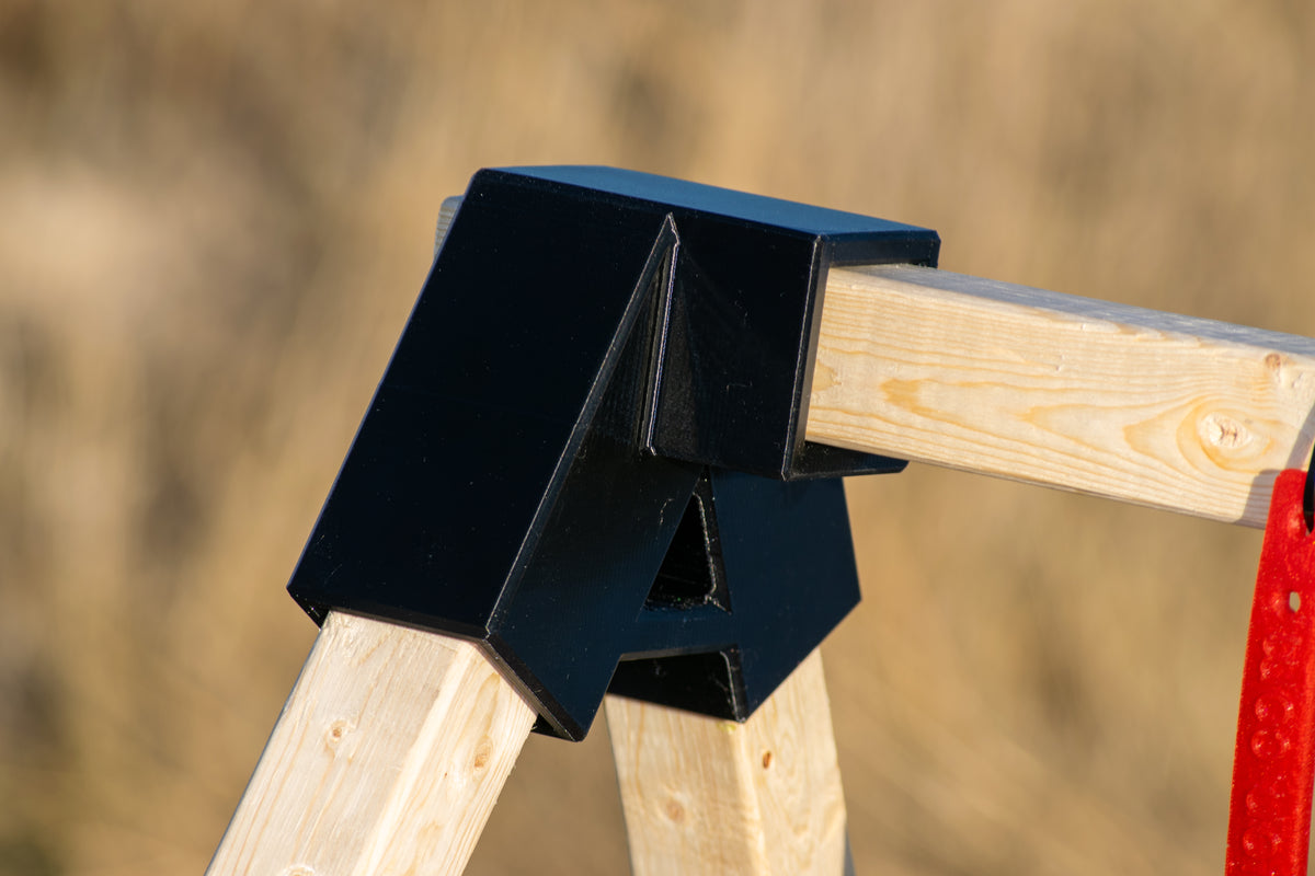 A close-up of a black plastic protective guard or bracket mounted on a light-colored wooden frame, with a red strap hanging on the right side against a blurred outdoor background.