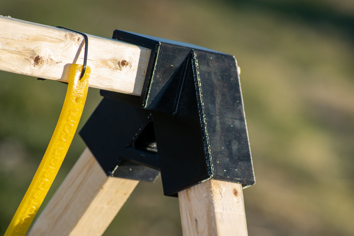 Close-up of a black plastic corner protector or bracket mounted on wooden beams outdoors, with a yellow hose hanging nearby against a blurred green background.