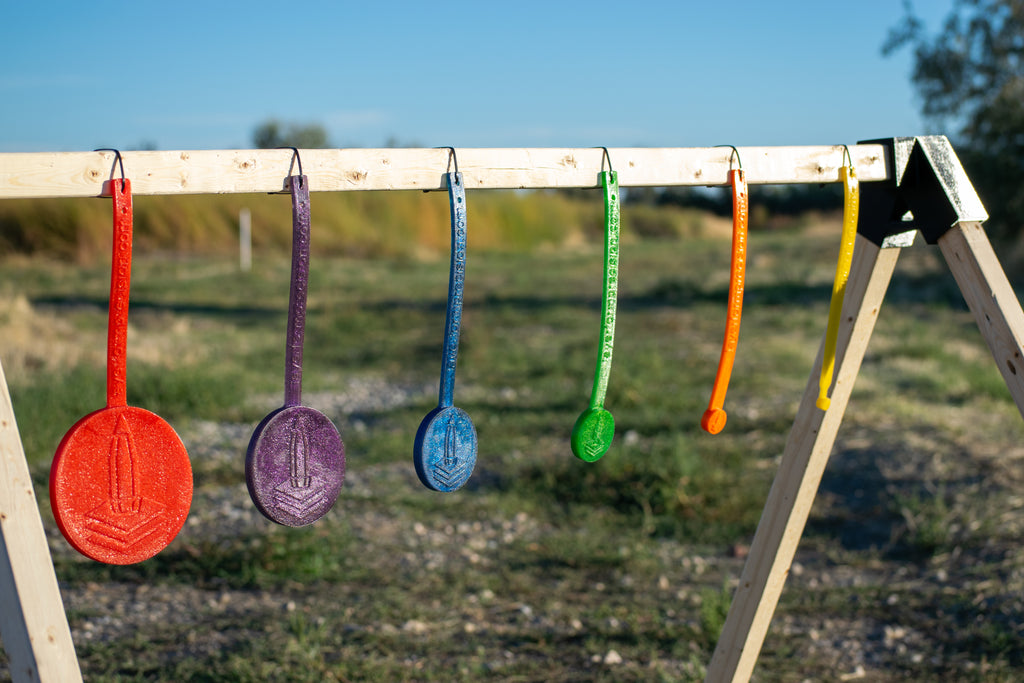 Five colorful circular hanging targets in red, purple, blue, green, and orange are suspended from a wooden frame outdoors on a grassy field under a blue sky.