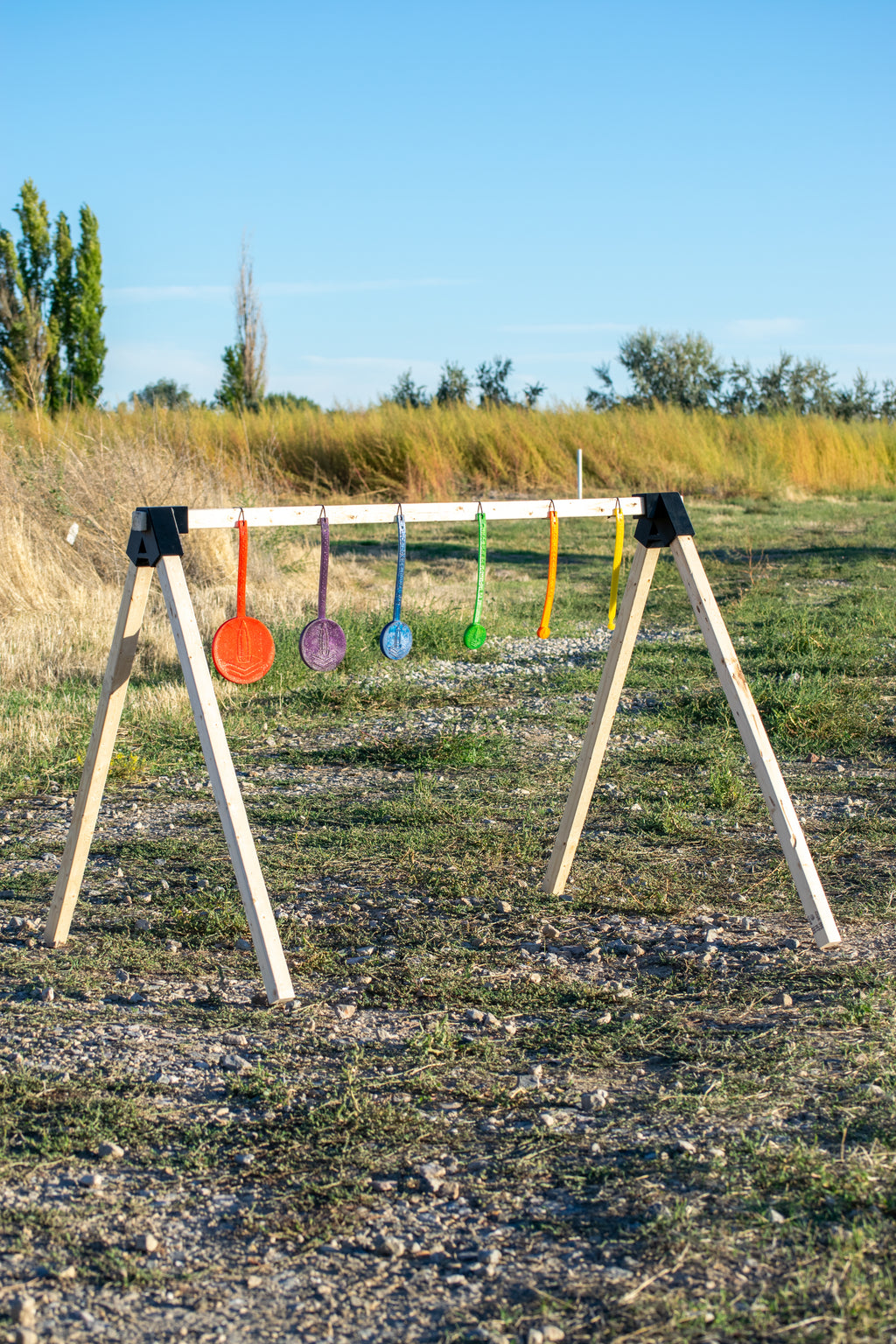 A wooden A-frame target stand set outdoors in a grassy field under a blue sky, holding six colorful hanging targets in red, orange, purple, blue, green, and yellow.