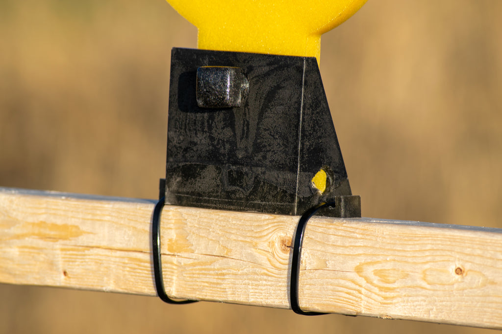 Close-up of a black plastic mounting bracket attached to a light wood post with black wire ties, with a yellow object partially visible at the top against a blurred tan background.