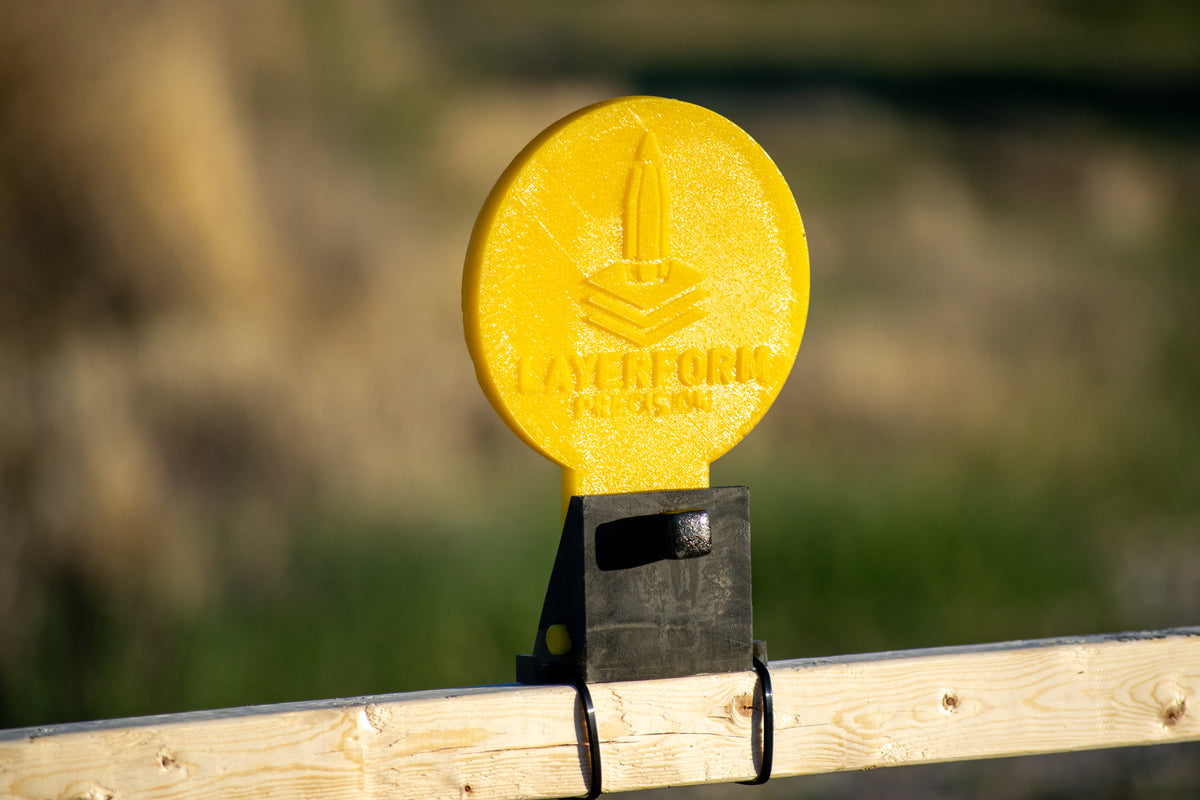 A yellow layerform impact target mounted on a black bracket attached to a wooden fence post, with a blurred outdoor background.