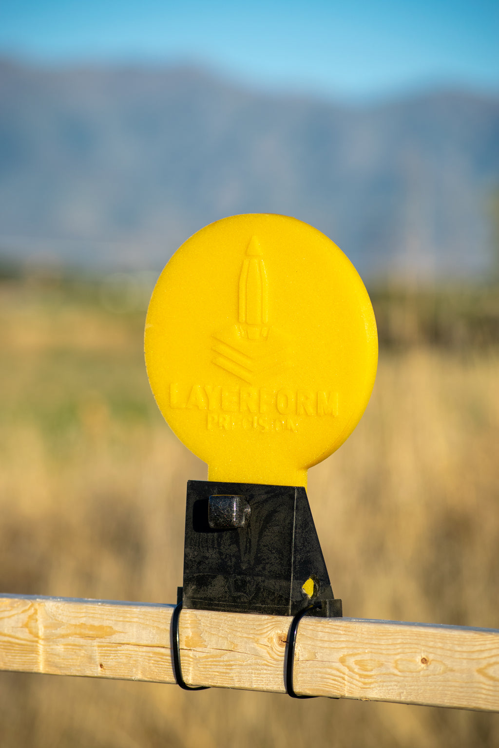A bright yellow circular layerform target with a black mounting bracket attached to a wooden fence post, set against a blurred outdoor background of dry grass and distant mountains.
