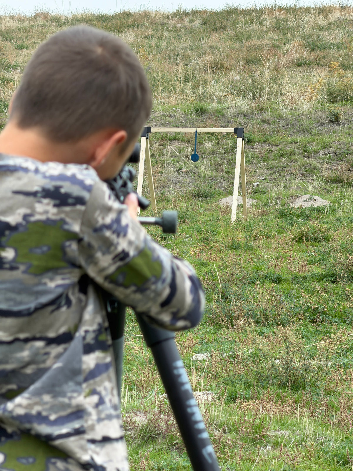 A person in a green and gray digital camouflage hunting jacket aims a rifle outdoors in a grassy field, with a target stand visible in the distance.